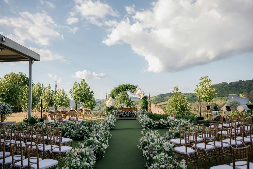 Covered terrace set for a wedding ceremony with white chairs, a floral arch and open sky beyond