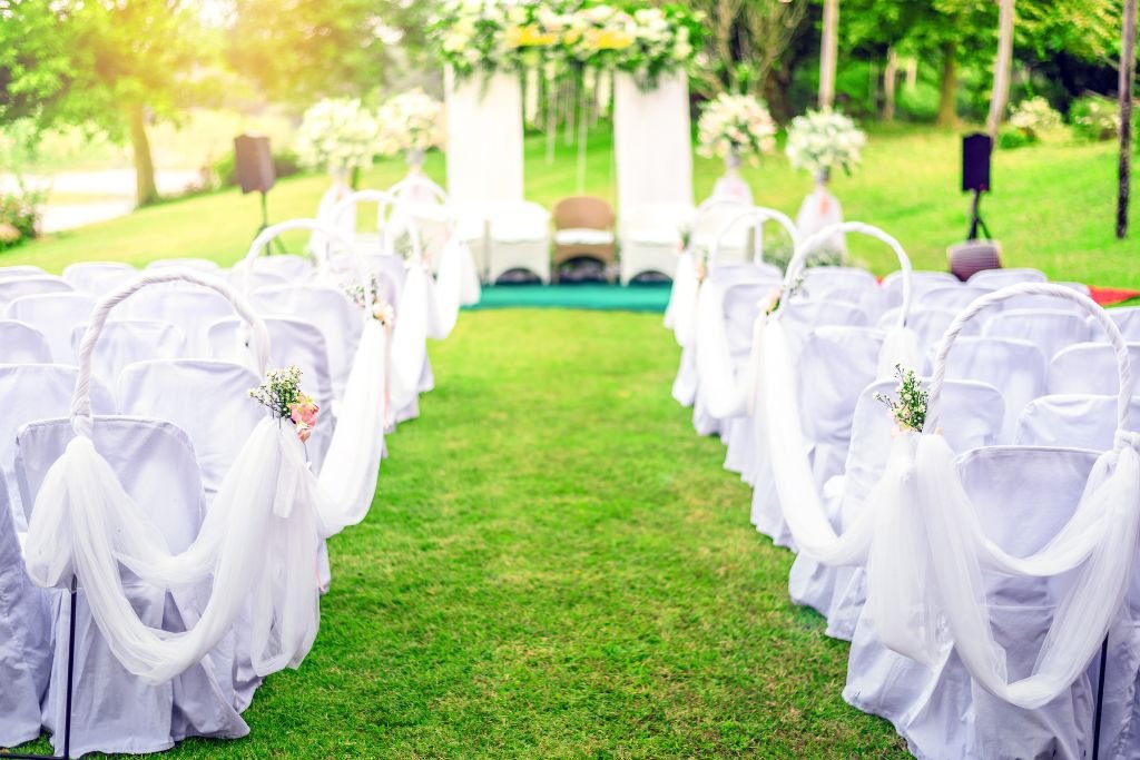 Outdoor wedding aisle on a lawn lined with white draped chairs and bows, leading to a decorated arch