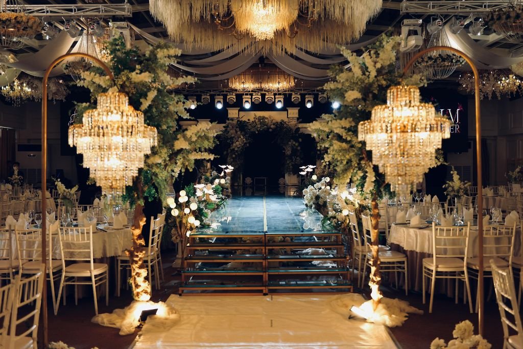 Candlelit ballroom set for a wedding ceremony, with crystal chandeliers above a long aisle and gold chiavari chairs