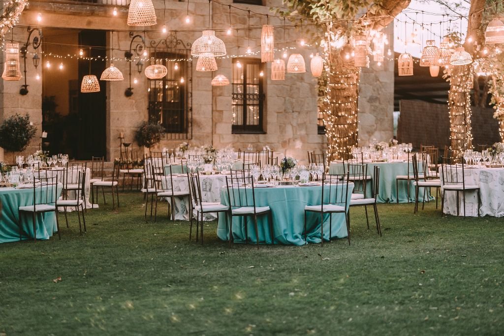 Exposed brick reception space set with round tables, teal linens and festoon lighting overhead