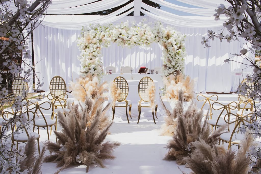 Draped white wedding ceremony backdrop with pampas grass, dried florals and cane chairs