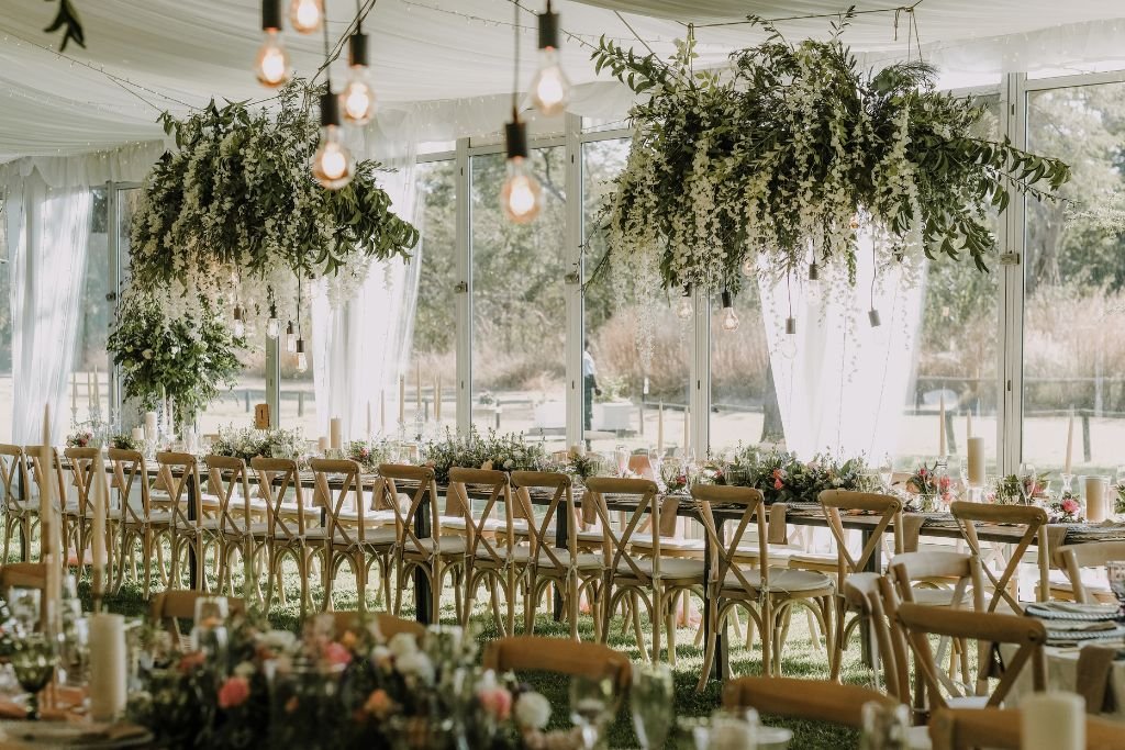 Wedding ceremony aisle under a canopy of hanging greenery and warm festoon bulbs, with cross back chairs