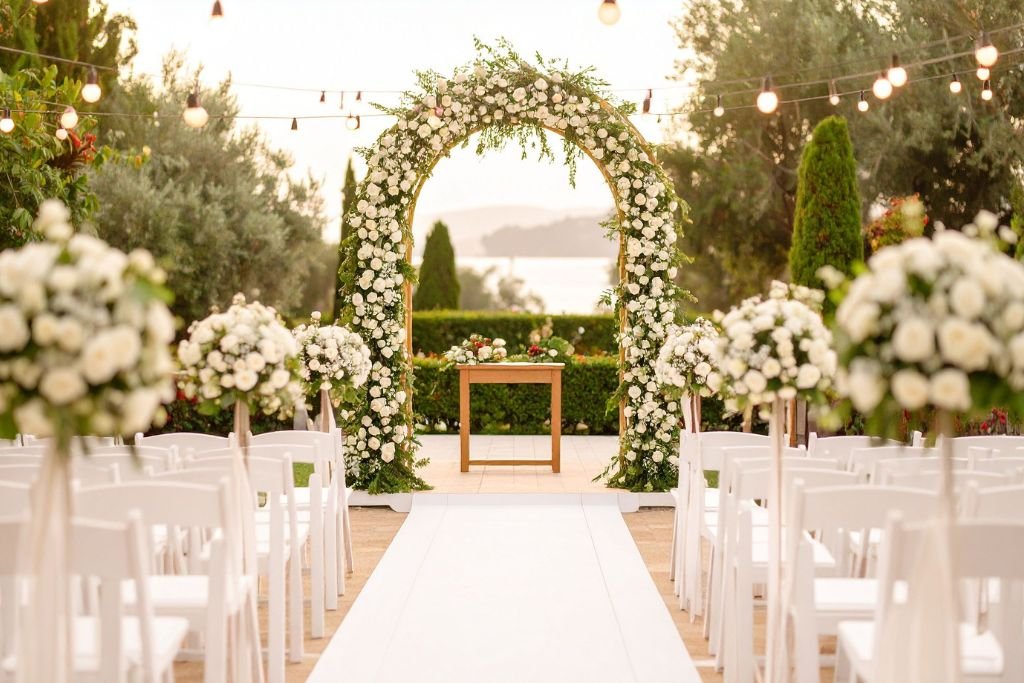 Wedding ceremony arch covered in white flowers at the end of a white aisle, with water and evening light behind