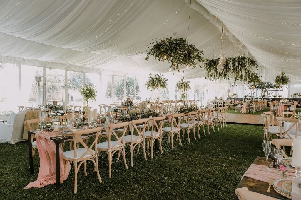Interior of a white marquee set for a wedding with cross back chairs, soft drapes and hanging greenery